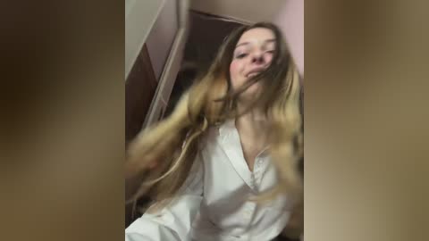 Video of a young Caucasian woman with long, blonde hair, wearing a white bathrobe, standing in a dimly lit bathroom with beige walls. Her face is partially blurred.
