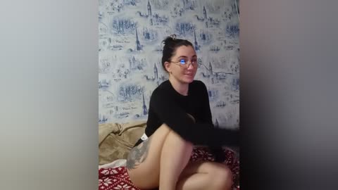 Video of a fair-skinned woman with dark hair in glasses, sitting on a bed with red and white patterned blanket, wearing black long-sleeve top and white shorts, against a blue and white wallpaper background.