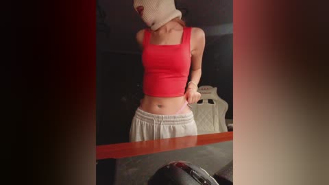A video of a slim, light-skinned woman with a red crop top and white sweatpants, wearing a beige beanie, standing indoors.