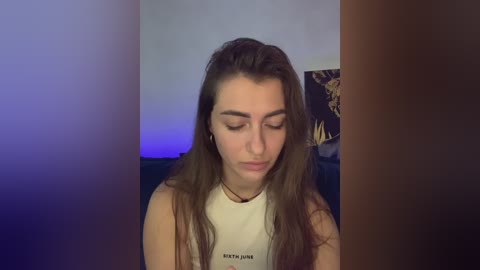 Video of a young woman with long brown hair, wearing a white tank top, sitting in a dimly lit room with a blue and purple light gradient.