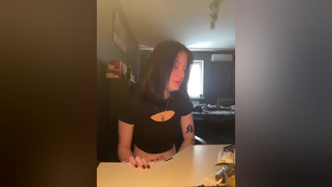 Video of a young woman with shoulder-length black hair, wearing a black T-shirt with a small design, leaning on a desk in a dimly lit room with dark furniture and a bed in the background.