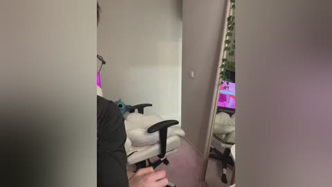 Video of a person wearing a black shirt, standing in a dimly lit room with beige walls and a white chair. A large mirror reflects a pink-lit gaming setup in the background.