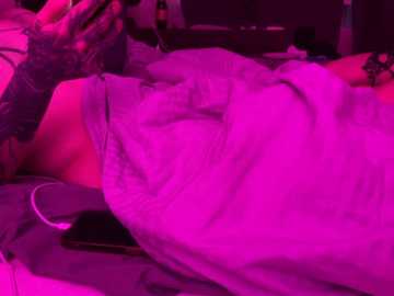 Video of a person lying on a bed, covered in a pink sheet, with a black phone in hand, bathed in a neon pink light.