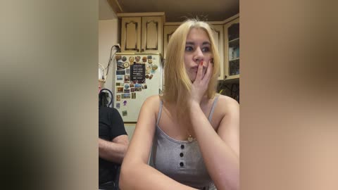 Video of a young blonde woman with fair skin and straight hair, wearing a gray tank top, sitting in a kitchen, covering her mouth with her hand. The kitchen has wooden cabinets, a refrigerator covered with magnets, and a man in a black shirt visible in the background.