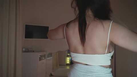 A video of a woman in a white ribbed tank top and white pants, viewed from behind, walking away in a dimly lit room with a flat-screen TV on the wall.