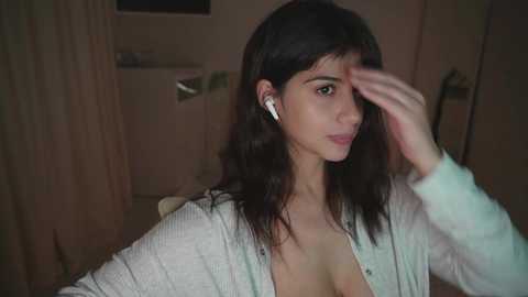 A video of a young woman with medium-length dark hair, wearing a white cardigan, headphones, and resting her hand on her forehead, sitting indoors with dim lighting.