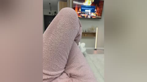 Video of a person's legs, clad in soft, pink pajama pants, reclining on a plush, white carpeted floor. In the background, a modern living room with a flat-screen TV showing a fireplace scene, and a tall, white floor lamp.