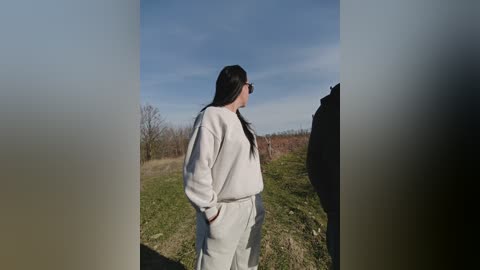 Video of a young woman with long black hair, wearing white sweatpants and a matching sweatshirt, standing outdoors in a grassy field under a clear blue sky, with a blurry foreground.