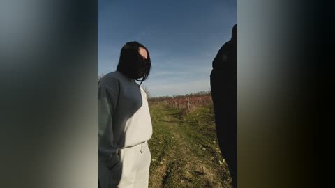 Video of a person wearing a light grey hoodie, standing in a barren, grassy field under a clear blue sky, with a blurred figure partially visible on the right.