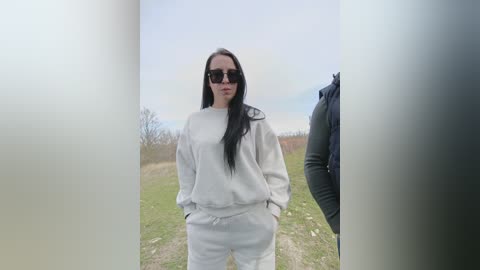 Video of a woman with long black hair, wearing large sunglasses and a white hoodie and pants, standing outdoors on a grassy path with a cloudy sky background.