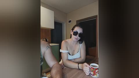Video of a woman with a dark complexion, long black hair, wearing a white off-shoulder top, sitting on a wooden floor, holding a red coffee mug, and a mirror partially obscuring the scene.