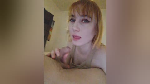 Video of a young woman with short, light brown hair and pale skin, wearing a beige top, leaning over a man's erect penis, in a dimly lit room with beige walls.