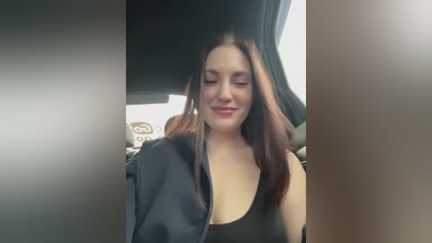 A video of a young woman with fair skin, long brown hair, and a slight smile, wearing a black jacket over a light-colored top, sitting in a car with a blurred interior background.