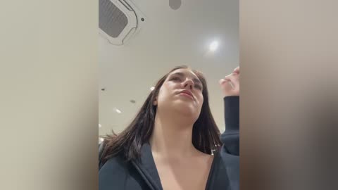 A video of a young woman with straight, dark brown hair, fair skin, and a black hoodie, standing in a dimly lit, beige-walled room with an air vent and a ceiling light.