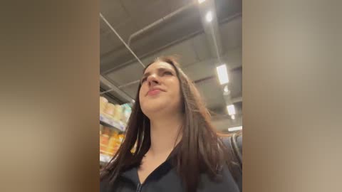Video of a young woman with long brown hair, wearing a black jacket, standing in an indoor space with shelves in the background.