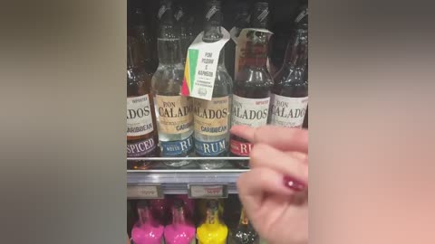 Video of a supermarket shelf displaying a variety of Dos Maderas rum bottles in glass bottles with different labels, prices, and flavors. A hand with a manicured nail holds a bottle.