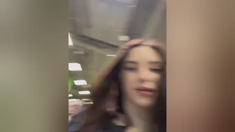 A blurry video of a young woman with long hair, eyes closed, touching her head, taken indoors with fluorescent lighting, possibly in an office setting.