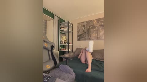 A video of a cozy bedroom with a large, gray gaming chair, a white person lying on a bed, a dark green bedspread, a beige wall, and a large abstract art piece.