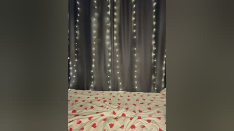 Video of a room with a white bedspread adorned with red heart patterns, flanked by dark gray curtains lit with string lights, creating a romantic, cozy atmosphere.
