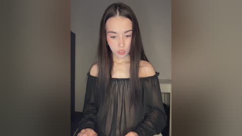 A video of a young Asian woman with long, straight black hair, wearing a sheer black off-shoulder top, standing in a dimly lit room.