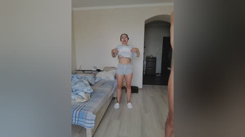 A video of a young woman in a bedroom, lifting her white crop top, revealing her midriff and small breasts. She wears light blue shorts and white sneakers. The room has a bed with plaid bedding, a dresser, and an open doorway.