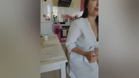 A video of a woman in a white, patterned bathrobe, standing in a modern, white kitchen with pink accents, holding a cup.