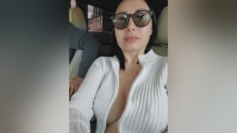 A video of a light-skinned woman with dark hair, wearing large sunglasses and a white ribbed knit sweater with a plunging neckline, sitting in a car.