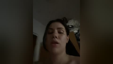 A dimly lit video of a woman with closed eyes, fair skin, and dark hair, possibly in a bedroom with a bed and clothes visible in the background.