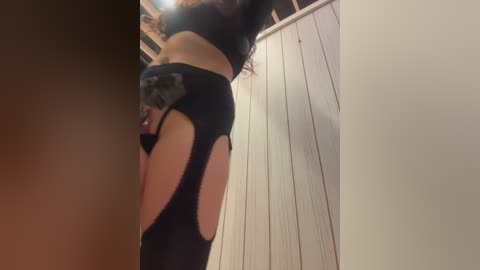 A video of a woman with light skin, wearing black lingerie with large cutouts, standing in a wooden-paneled room.