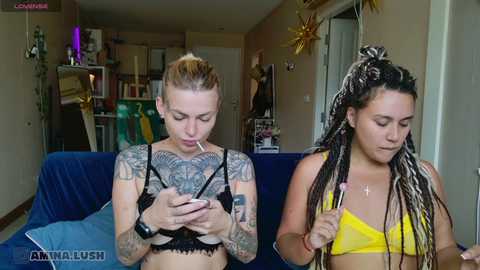 Video of two tattooed women with light skin, one in a black bra, the other in a yellow bikini, sitting on a blue couch in a cozy living room.