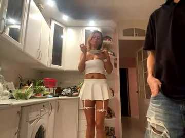 Video of a blonde woman in a revealing white mini-skirt and crop top, holding a phone in a modern kitchen with white cabinets and a man in jeans.