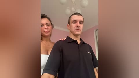 A video of a man and a woman standing closely together in a room with a pink wall and white ceiling light. The woman, with a white dress, has her arm around the man's shoulders. He wears a black polo shirt.