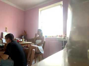 A video of a dimly lit room with pink walls, showing a young woman in a white dress and a man in a black shirt, both seated at a desk, possibly in a classroom setting.