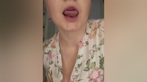 A video of a fair-skinned woman with full lips sticking out her tongue. She wears a floral-patterned blouse. The background is blurred, showing a beige wall.