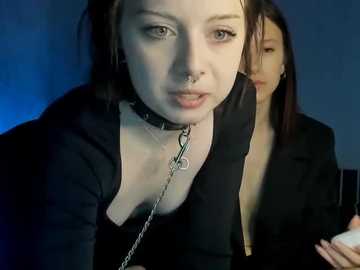 Video of a young, pale-skinned woman with dark hair and piercings, wearing a black leather collar and chain, in a dimly lit room, with another person partially visible in the background.