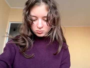 A video of a young Caucasian girl with long, wavy brown hair, wearing a purple turtleneck sweater, looking down thoughtfully in a beige room with a white door in the background.