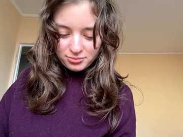 Video of a young girl with long, wavy brown hair, wearing a purple sweater, standing indoors against a beige wall. She has a soft, gentle expression and slightly closed eyes.