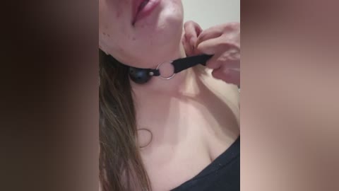 Video of a young woman with light skin, brown hair, wearing a black leather choker with a metal ring, holding her collar with one hand. Background blurred.