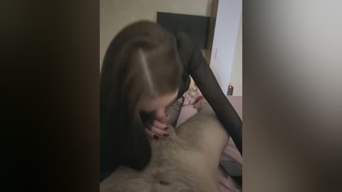 A video of a woman with long brown hair, dressed in black, performing oral sex on a man with light brown pubic hair. The setting is a dimly lit bedroom with a bed and neutral-toned walls.