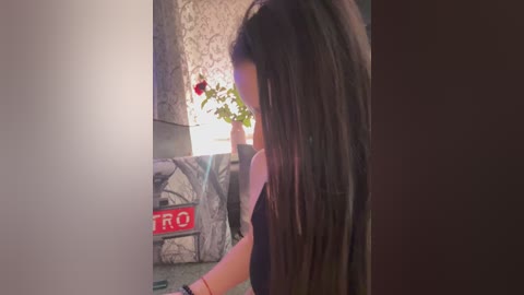 A video captures a young woman with long, straight brown hair, wearing a black top, leaning against a wall adorned with a red \"STOP\" sign and a potted plant.