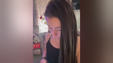 Video of a young woman with long black hair, wearing a black tank top, looking down at her phone. Background features a red \"DRO\" sign and a floral arrangement.