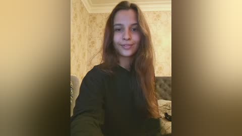 Video of a young woman with long brown hair, wearing a black sweater, seated on a chair in a dimly lit bedroom with beige wallpaper and a tufted headboard.