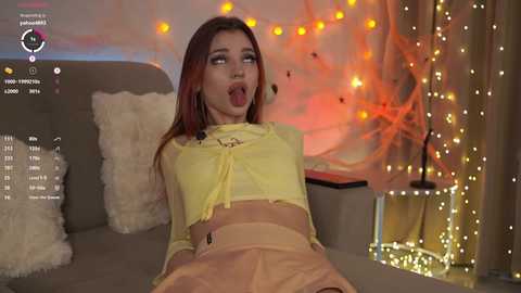 Video of a young woman with fair skin and long red hair, wearing a yellow crop top and high-waisted skirt, posing suggestively on a couch in a cozy, warmly lit room with string lights and soft textures.
