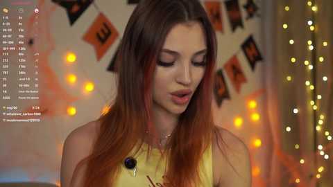 Video of a young woman with long, straight, reddish-brown hair, wearing a yellow sleeveless top, smiling softly, standing in a warmly lit, festive room with string lights and Halloween decorations in the background.