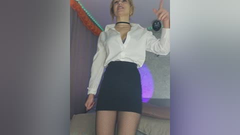Video of a blonde woman in a white blouse, black skirt, fishnet stockings, and a choker, posing provocatively with a remote control in a dimly lit room with neon lights.