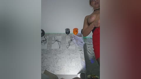 Video of a shirtless, topless man with a chiseled physique, wearing red pants, standing in a dimly lit room with a white marble wall adorned with black and orange abstract art, partially obscured by a red curtain.