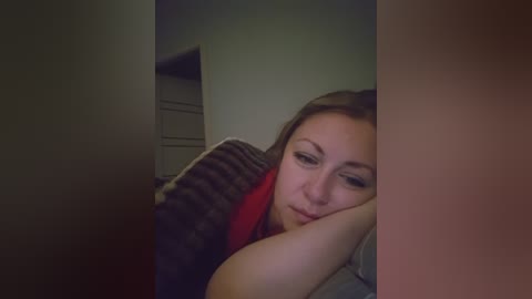 A video of a young woman with light skin, light brown hair, and wearing a red top, lying on her side in a dimly lit room, with a blurry, dark object partially obstructing the background.