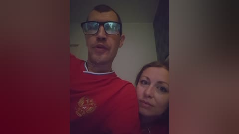 Video of a man with glasses and a red shirt featuring a gold emblem, standing next to a woman with long hair, both indoors.