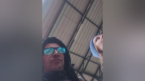 Video of a man with a serious expression, wearing black sunglasses and a dark hoodie, standing in a dimly lit, corrugated metal-roofed building. The background is blurry.