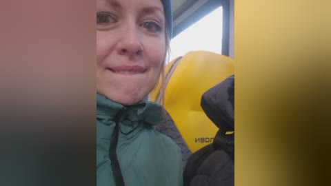 Video of a smiling woman with fair skin and blue eyes, wearing a green puffer jacket, holding a yellow and black climbing helmet. She is inside a vehicle with a window view.
